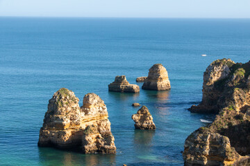 Fototapeta premium Coastal rock formations rise from calm blue waters under clear skies in Algarve, South of Portugal