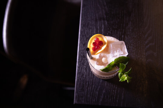 Elegant cocktail with ice, mint, and a lemon garnish topped with red berries, served with a black straw on a dark wooden table