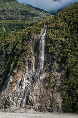 Dramatic tall waterfall after heavy rain at Franz Josef Glacier surrounded with green nature with mist hovering in the background
