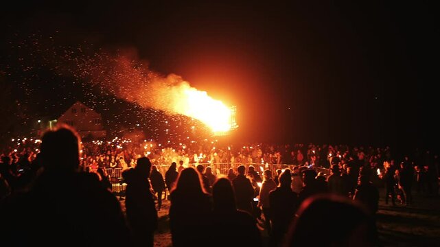A large crowd of people are gathered around a fire, with some holding lit candles. The atmosphere is lively and festive, with people enjoying the warmth and light of the fire