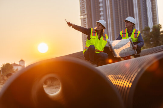 two construction workers, one kneeling and the other standing, on large pipes at a construction site. They are reviewing blueprints and discussing a pipe system for the project
