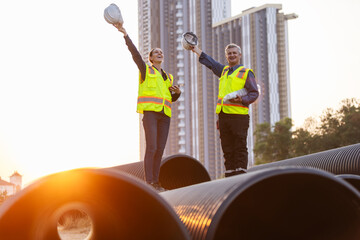 Two engineers stand on large water pipes at a construction site, celebrating the completion of...