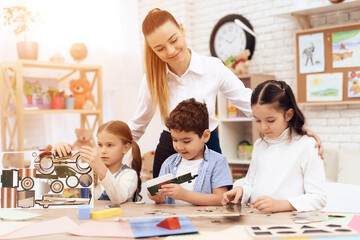 Fototapeta premium A young woman helps children fold parts from cardboard. This is their teacher. They have a lesson.
