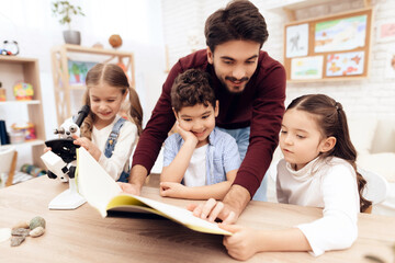 The children together are reading the book together. They have a lesson. An adult teacher helps them.