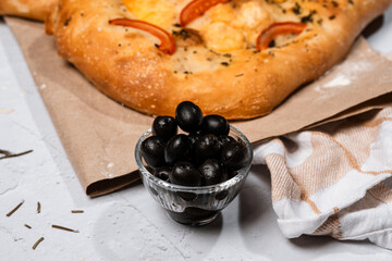 focaccia with cheese, olives, olive oil, tomatoes on kraft paper on a light background, next to a cheese grater, a bowl of olives, Italian herbs, Italian pastries, yeast dough