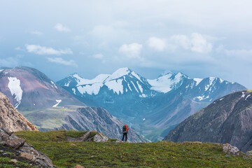 Man in vivid red jacket admire alpine scenery on green grassy hill among rocks near precipice edge. Guy with backpack on pass enjoying few big snowy pointy peaks. Three large snow peaked tops far away