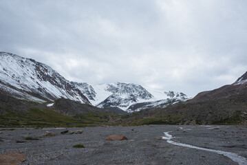 Dramatic cold landscape with alpine river on stony glade against snow covered large mountain range under rainy cloudy gray sky. Gloomy rainy weather in high mountains. Big snowy mount in rain time.
