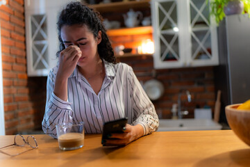 Frustrated businesswoman rubbing her nose bridge while reading upsetting news on her smartphone, sitting at the kitchen table with a cup of coffee and eyeglasses next to her