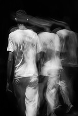 The blurred motion of people walking is shot in black and white. The composition focuses on their blurred shapes against an isolated background, emphasizing modern fashion with sharp lines and strong 