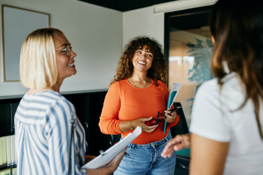 Friendly Office Interaction Among Diverse Women in a Modern Workspace. The three women are talking together, smiling, with notebooks in their arms