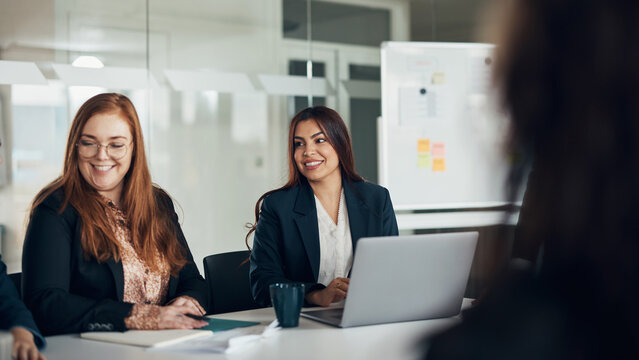 Smiling diverse businesswomen sitting at a conference table during an office meeting. There is a whiteboard in the background with post it notes on it.