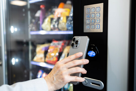 Businesswoman buying snacks on vending machine on job break and making contactless payment via smart phone.