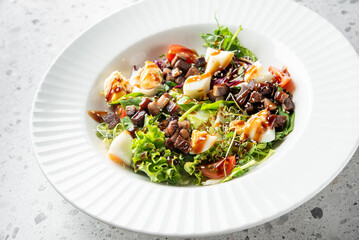 A fresh salad in a white bowl featuring mixed greens, diced meat, boiled eggs, and cherry tomatoes, drizzled with a savory sauce, presented on a textured gray surface