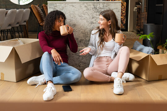 Sitting on floor with unpacked boxes, lesbian couple drinking coffee and chatting - Powered by Adobe