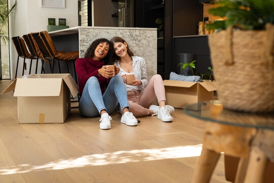 Lesbian couple sits on floor with unpacked boxes, enjoying coffee in their new home - Powered by Adobe