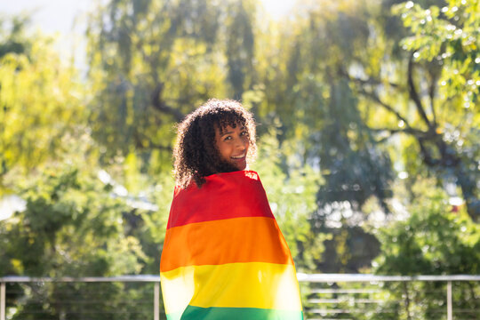 Smiling woman wrapped in rainbow flag standing outdoors, enjoying sunny day