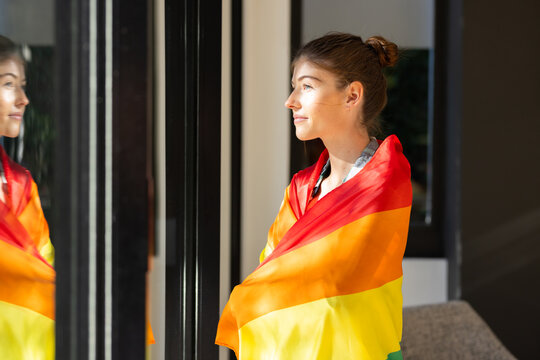 Woman wrapped in rainbow flag standing by window, reflecting in glass