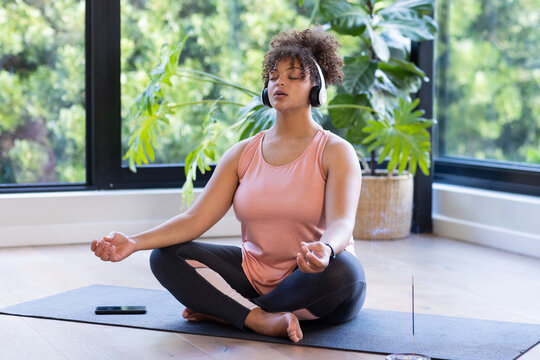 Meditating on yoga mat, plus size woman wearing headphones in peaceful home setting, at home, at hom