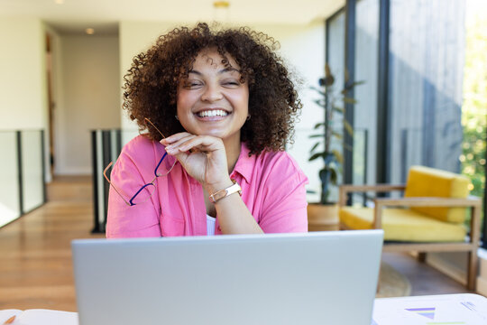 Smiling woman holding glasses and working on laptop in modern home office, at home