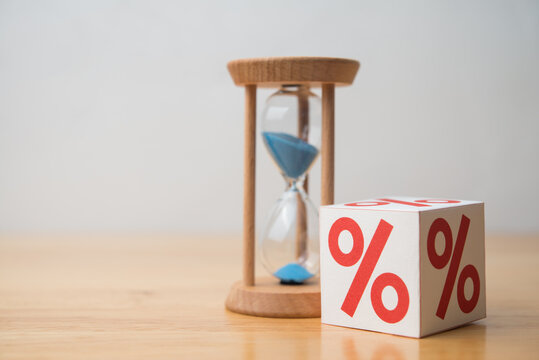 Percent (or Interest rate) sign and hourglass on wooden table with white wall background copy space. Interest is a cost of doing business. Business, finance and economy concept.