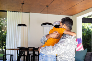 Hugging his young son, soldier in uniform holding American flag indoors, copy space