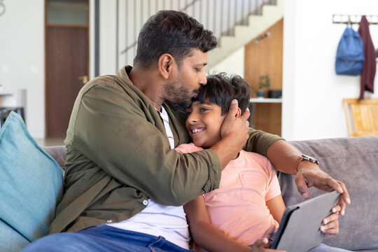 indian father hugging young son holding tablet, sharing loving moment on couch