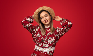 Positive young female in trendy floral dress and straw hat smiling while looking away standing...