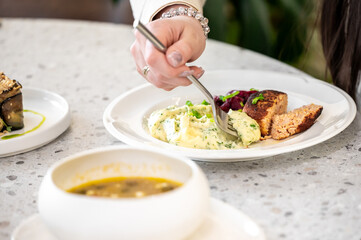 A close-up of a hand holding a fork, enjoying a plate of creamy mashed potatoes, vibrant salad, and a slice of salmon cake on a textured table, with a bowl of soup in the background