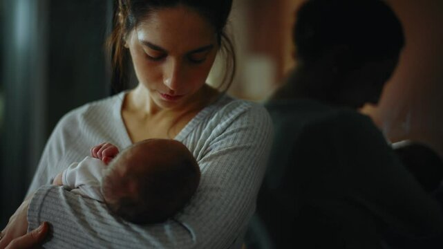 Authentic footage of a mother in pajamas at night trying to put her newborn baby to sleep in her arms. The mother is very tired and yawns and cuddles her baby in the bedroom