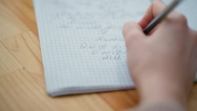 Close-up hand view of person writing on wooden table with black pen, cursive handwriting visible on notepad, focused penmanship with soft natural lighting and blurred background