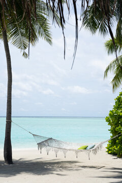 A white hammock tied to a palm tree on a sandy beach, overlooking turquoise waters and a tropical island. A perfect spot for relaxation in paradise in Maldives
