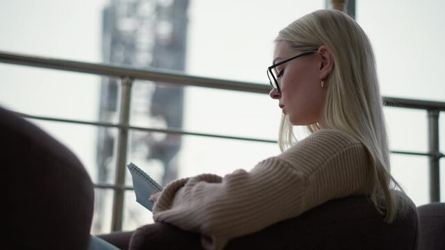 Young lady seated, focused and writing on her journal with a thoughtful expression, wearing glasses, in a cozy indoor environment with natural light streaming through a glass panel