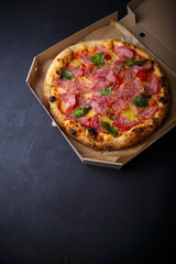 Pizza with cheese, ham, green basil leaves and tomatoes in paper box on black background. Food product advertising banner, copy space