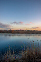 Obraz premium Beautiful blue hour on the lake, trees on the beach , sunrise over the lake with blue sky without clouds.Landscape photography on the pond , purple colors on the sky , trees and water , reflections 