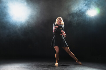 Fototapeta premium Contemporary woman dancer posing on black background with smoke. Adult female artist performer dancing with expression. Modern ballerina dancing in darkness. Passion dance in dark. Flexibility balance