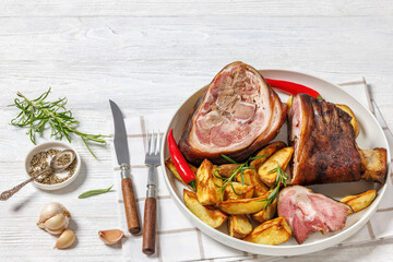 Smoked pork knuckle, ham hock with roasted potatoes, red chili peppers on whie platter on white wooden table with cutlery, horizontal view