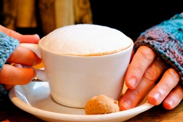 Close-up female hand in blue gloves holding cappuccino and cafe latte. Hot drinks in winter. Cafe culture. Female hand holding hot coffee. Cold weather, knitted glove. Coffee with heart.