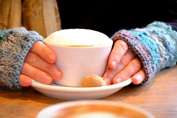 Close-up female hand in blue gloves holding cappuccino and cafe latte. Hot drinks in winter. Cafe culture. Female hand holding hot coffee. Cold weather, knitted glove. Coffee with heart.