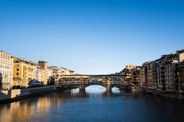 Front view of the Arno River with the Ponte Vecchio bridge in the background. The dawn light illuminates the facades that give the river and are reflected in the water in a clear day