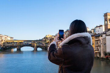 A young woman takes a photo with her phone of the Ponte Vecchio bridge in the background. The dawn light illuminates the facades that give the river and are reflected in the water in a clear day © LaMorenita