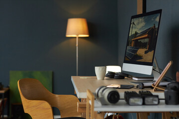 Photography equipment placed on organized wooden desk beside modern computer in cozy room. Soft lighting provided by standing lamp enhances warm ambiance
