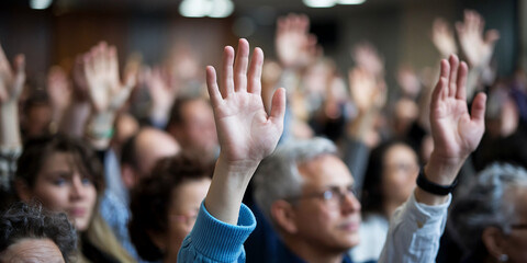 A diverse group of individuals raising their hands in a crowd, participating in a public event or meeting, symbolizing engagement, agreement, or decision-making.