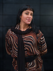 A confident woman in a stylish brown patterned blouse poses playfully against a sleek dark background, showcasing modern fashion and natural charm in an urban setting
