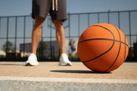 Basketball orange ball lies and athletes legs at outdoor public basketball court. Sports gear. Template, sport background. Close up. Outdoors.