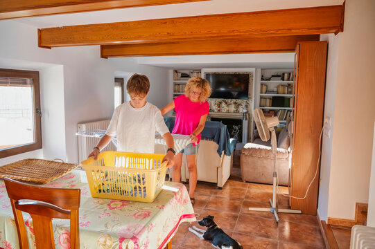 Middle-aged woman and her teenage son doing household chores together at home, folding clothes and organizing laundry in a cozy living room with a dog resting on the floor