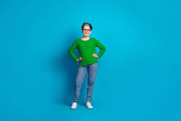Confident elderly woman poses in green sweater and jeans on vibrant blue background