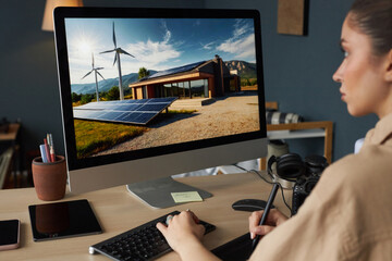 Individual working on design of renewable energy system at computer with wind turbines and solar panels displayed on screen. Modern workspace including various digital devices and tools