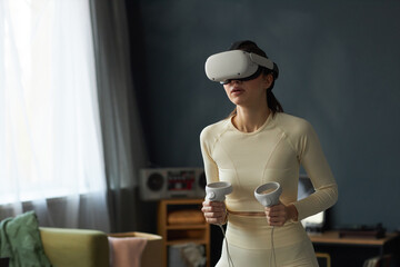 Individual standing in living room, using virtual reality headset and holding controllers,...