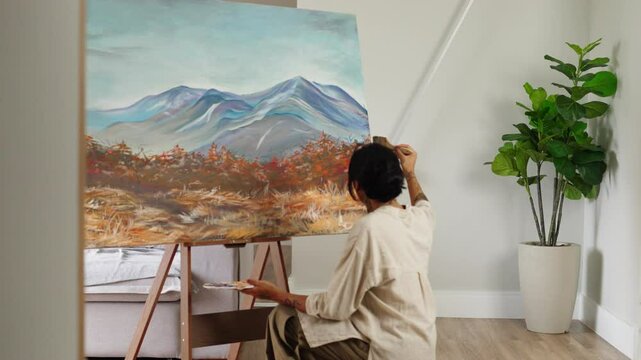 An artist applies colorful acrylic paint to a canvas with a brush. A young woman artist works in her workshop. Nature painting, contemporary art.
