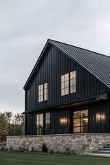Beautiful modern black farmhouse with stone foundation and large windows located in a scenic rural area surrounded by greenery on a cloudy day